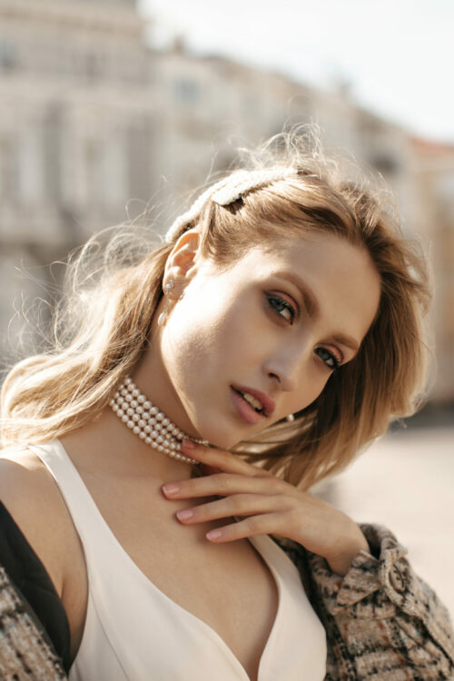 Close-up portrait of blonde gorgeous woman in beautiful makeup looking into camera outside. Attractive lady in white dress and checkered jacket poses at city square.