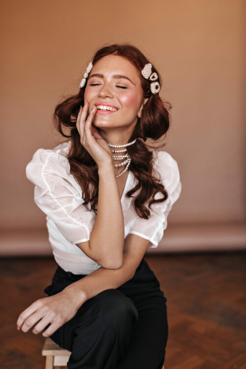 Beautiful curly girl dressed in white blouse and black pants sits on chair. Woman with hairpins and pearl necklace posing in beige room