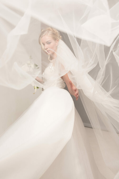 Bride in beautiful dress standing indoors in white studio interior like at home. Trendy wedding style shot.